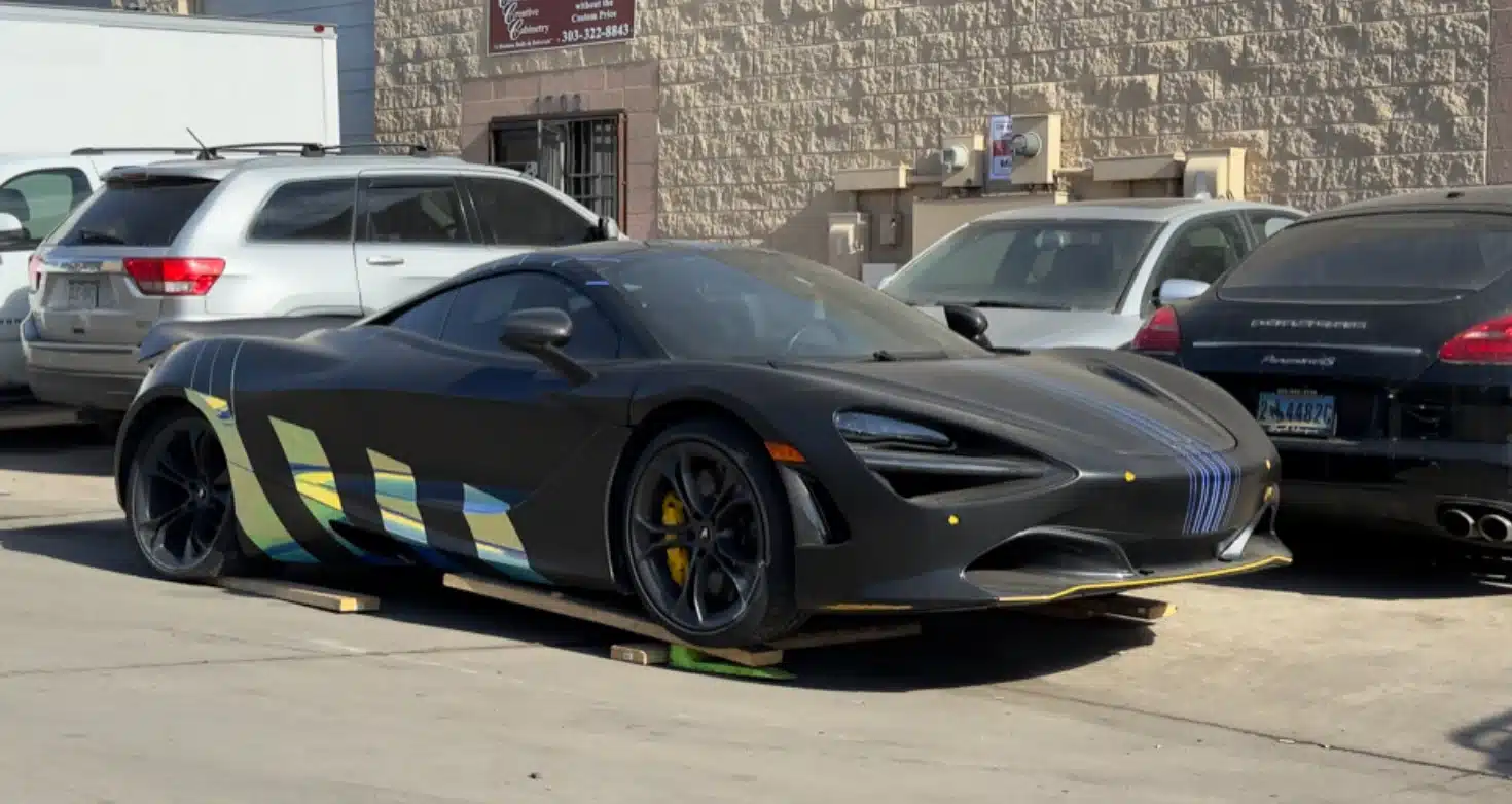 A matte black McLaren 720S with custom blue and yellow graphics parked on wooden planks outside an auto repair shop, surrounded by SUVs and luxury cars.