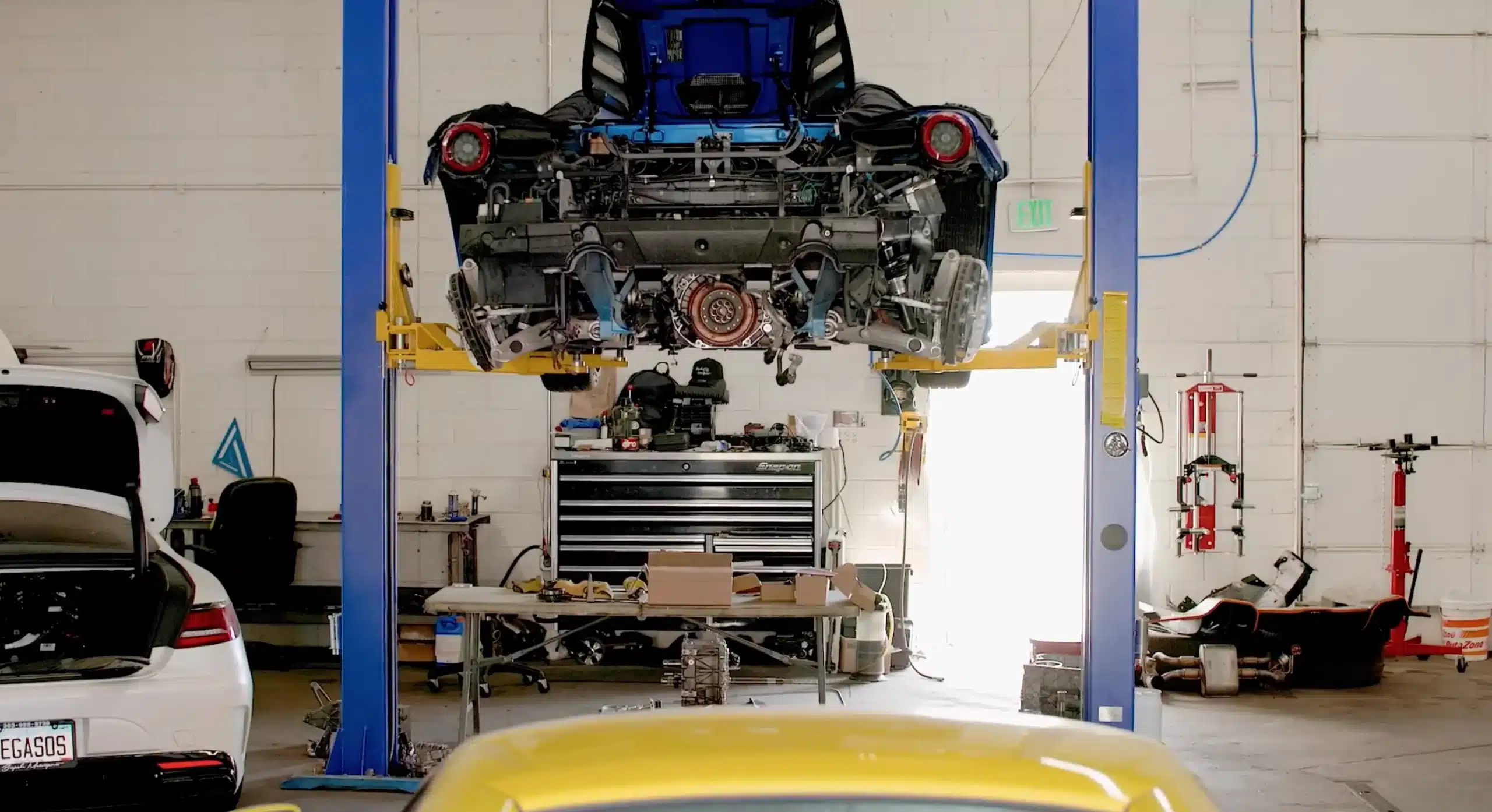 A blue Ferrari 488 Pista suspended on a hydraulic lift inside an auto repair shop, with its rear drivetrain and engine removed. Various tools, car parts, and a workbench with boxes and equipment are visible in the background.