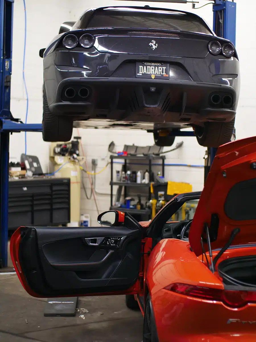 A black Ferrari on a hydraulic lift in a repair shop, with an orange convertible sports car in the foreground having its door and trunk open.