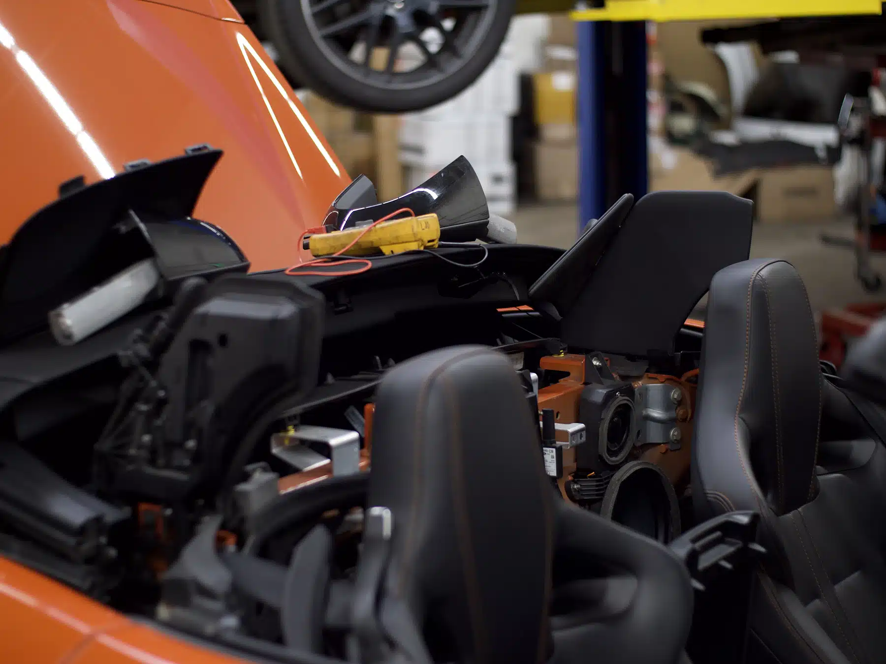Partially disassembled orange convertible car in a repair shop, exposing internal components and wiring, with tools placed on the dashboard.