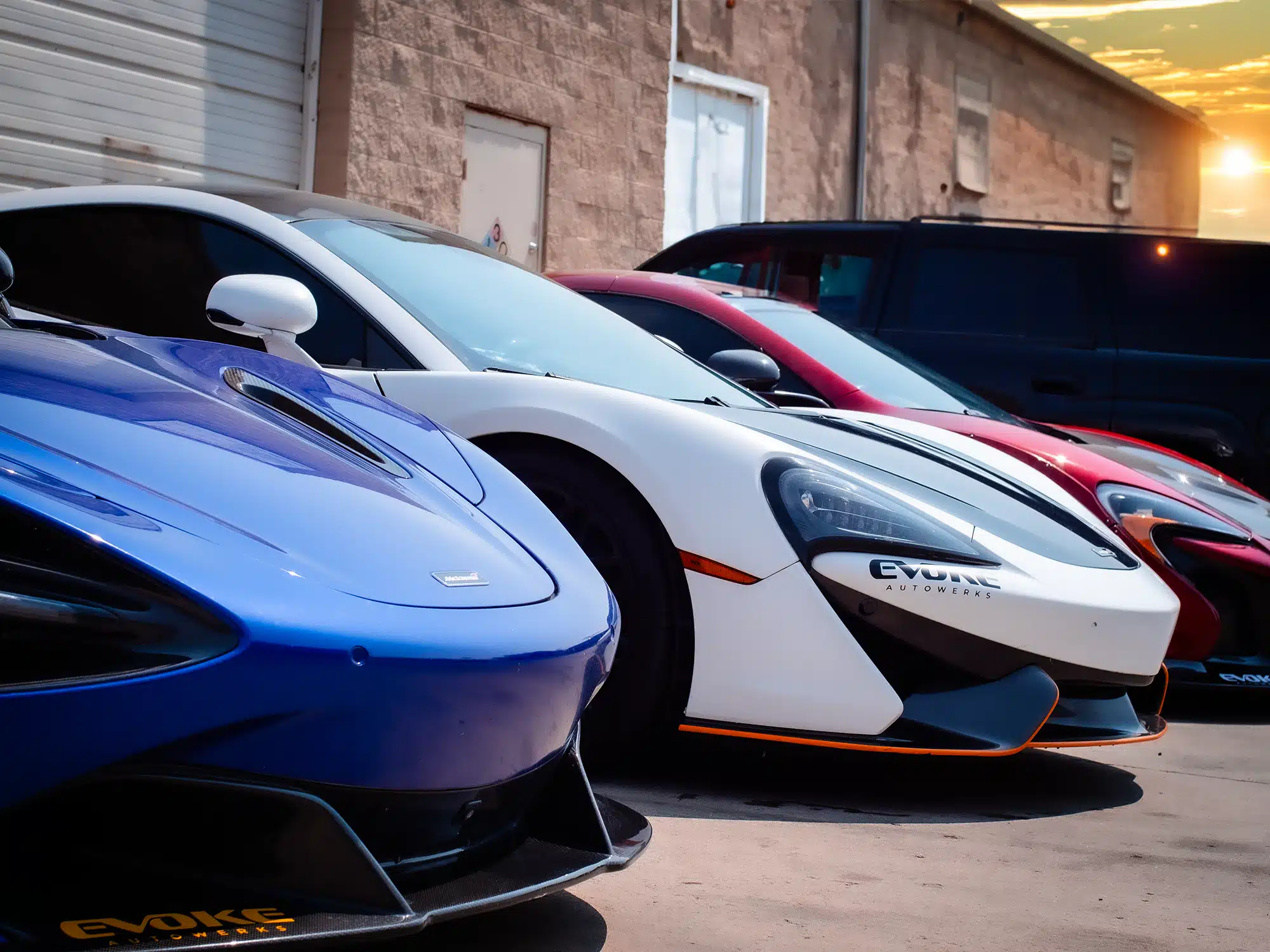Lineup of customized McLaren supercars, including blue, white, and red models with Evoke Autowerks branding, parked outside the workshop in Englewood, CO, at sunset.